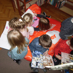 W bibliotece. Grupa dzieci siedzi na dużej pomarańczowej pufie i przeglądają książki.