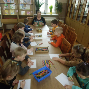 W bibliotece. Grupa dzieci siedzi przy długim stoliku i wykonuje prace plastyczne.