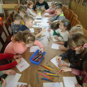 W bibliotece. Grupa dzieci siedzi przy długim stoliku i wykonuje prace plastyczne.