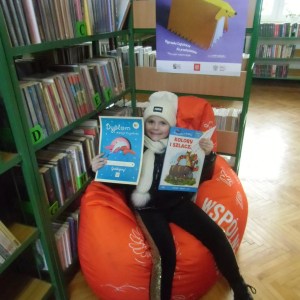 W pomieszczeniu biblioteki. Uśmiechnięta dziewczynka siedzi na pomarańczowej pufie. W ręku trzyma książkę i dyplom. W tle regały z książkami.