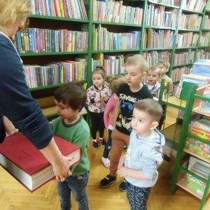 W bibliotece. Bibliotekarka trzyma wielką książkę, którą jedno z dzieci próbuje unieść.