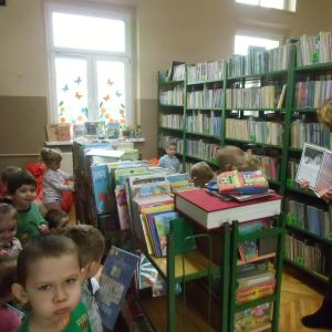 W bibliotece. Bibliotekarka trzyma w ręku książkę. Troje z dzieci przygląda się książce, reszta przegląda książki na regałach.