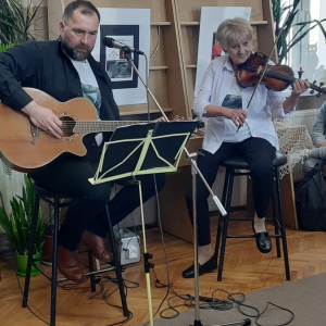 Koncert duetu. Mężczyzna siedzi na krześle i gra na gitarze. Obok niego na krzesle siedzi kobieta i gra na skrzypcach.