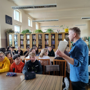 W bibliotece. Grupa dzieci siedzi przy stolikach. Prowadzący spotkanie pokazuje książkę