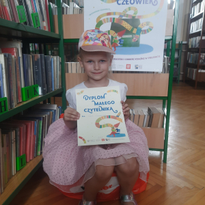 W pomieszczeniu biblioteki. Uśmiechnięta dziewczynka siedzi na pomarańczowej pufie. W ręku trzyma dyplom. W tle regały z książkami.