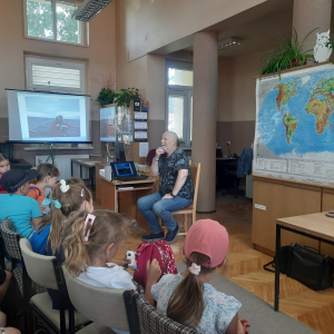 W bibliotece. Mężczyzna siedzi na krześle. Za nim znajduje się mapa świata. Obok, po lewej stronie znajduje się ekran, na którym jest zdjęcie z podróży prowadzącego spotkanie mężczyzyny.