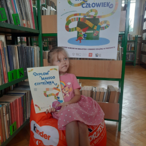 W pomieszczeniu biblioteki. Dziewczynka siedzi na pomarańczowej pufie.  W ręku trzyma dyplom. W tle regały z książkami.