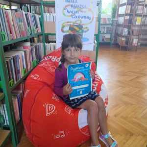 W pomieszczeniu biblioteki. Dziewczynka siedzi na pomarańczowej pufie. W ręku trzyma dyplom. W tle regały z książkami.