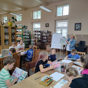 W bibliotece. Grupa dzieci siedzi przy stolikach. Patrzą w stronę kobiety, która prowadzi spotkanie i pokazuje komiksy.