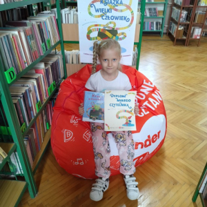 W pomieszczeniu biblioteki. Dziewczynka siedzi na pomarańczowej pufie. Trzyma książkę i dyplom. W tle regały z książkami.