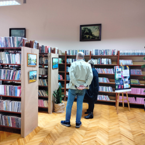 Wystawa obrazów w bibliotece. Na regałach przykrytych szarym papierem zawieszone są obrazy. Mężczyzna i kobieta oglądają wystawę.