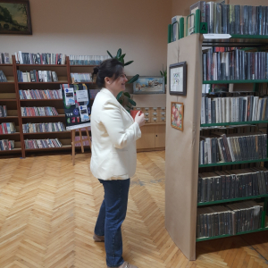 W bibliotece. Na regałach przykrytych szarym papierem zawieszone są obrazy. Kobieta uśmiecha się spogląda na obraz. Ubrana jest w jasną marynarkę i spodnie dżinsowe.