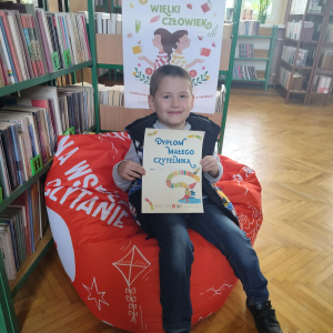 W pomieszczeniu biblioteki. Uśmiechnięty chłopiec siedzi na pomarańczowej pufie. W ręku trzyma dyplom. W tle regały z książkami.