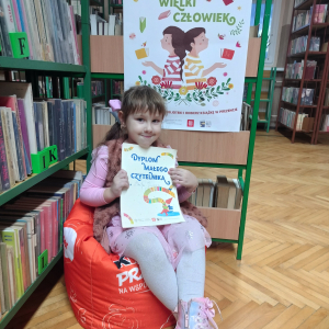 W pomieszczeniu biblioteki. Dziewczynka siedzi na pomarańczowej pufie. W ręku trzyma dyplom. W tle regały z książkami.