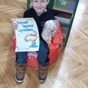 W pomieszczeniu biblioteki. Uśmiechnięty chłopiec siedzi na pomarańczowej pufie. W ręku trzyma dyplom i pluszowego pieska. W tle regały z książkami.