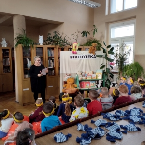 W bibliotece. Dzieci patrzą na kobietę, która prowadzi spotkanie. Obok niej znajduje się kotara, na której jest napisane biblioteka. Nad nią znajdują się kukiełki: lalka i miś.