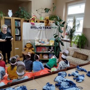 W bibliotece. Dzieci patrzą na kobietę, która prowadzi spotkanie. Obok niej znajduje się kotara, na której jest napisane biblioteka. Nad nią znajdują się kukiełki: lalka i miś.