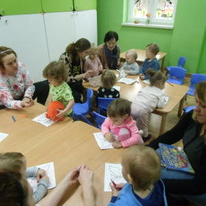 W budynku. Sala pomalowana na zielono. Dzieci siedzą przy stolikach i kolorują motyle. Pomagają...