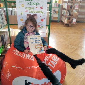 W pomieszczeniu biblioteki. Dziewczynka siedzi na pomarańczowej pufie. W ręku trzyma dyplom. W tle regały z książkami.