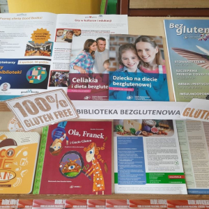 Wystawa książek i broszur otrzymanych w ramach nagrody za udział w konkursie "Biblioteka bezglutenowa". Książki i broszurki ustawione jedna obok drugiej, przodem okładki. Na środku wydrukowany napis Biblioteka bezglutenowa i 100% gluten free