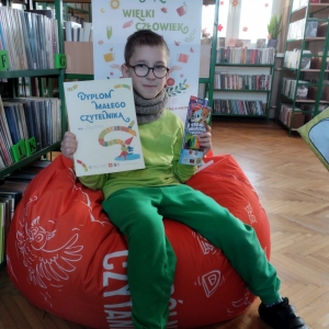 "W pomieszczeniu biblioteki. Chłopiec siedzi na pomarańczowej pufie. W ręku trzyma dyplom i opakowanie kredek. W tle regały z książkami