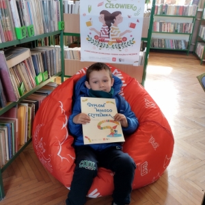 W pomieszczeniu biblioteki. Chłopiec siedzi na pomarańczowej pufie. W ręku trzyma dyplom. W tle regały z książkami.