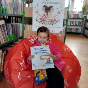 W pomieszczeniu biblioteki. Dziewczynka siedzi na pomarańczowej pufie. W ręku trzyma dyplom. W tle regały z książkami.