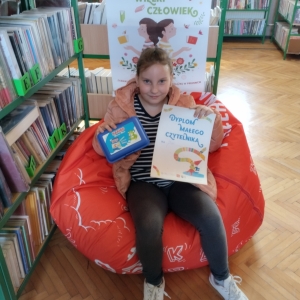 W pomieszczeniu biblioteki. Uśmiechnięta dziewczynka siedzi na pomarańczowej pufie. W ręku trzyma dyplom i niebieskie pudełko. W tle regały z książkami.