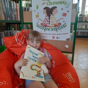 W pomieszczeniu biblioteki. Uśmiechnięta dziewczynka siedzi na pomarańczowej pufie. W ręku trzyma dyplom. W tle regały z książkami.