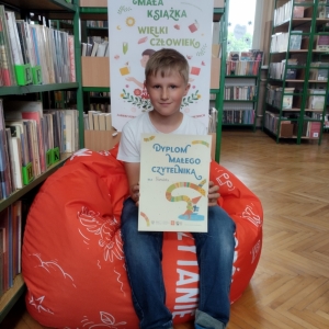 W pomieszczeniu biblioteki. Chłopiec siedzi na pomarańczowej pufie. W ręku trzyma dyplom. W tle regały z książkami.