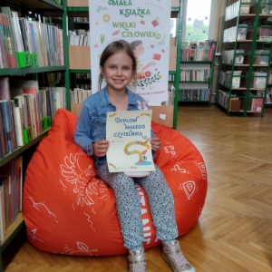 W pomieszczeniu biblioteki. Uśmiechnięta dziewczynka siedzi na pomarańczowej pufie. W ręku trzyma dyplom. W tle regały z książkami.