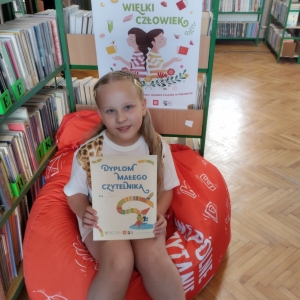 W pomieszczeniu biblioteki. Uśmiechnięta dziewczynka siedzi na pomarańczowej pufie. W ręku trzyma dyplom. W tle regały z książkami.