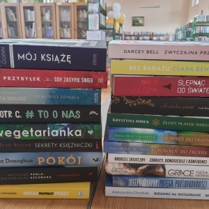 W bibliotece. Na stoliku ułożone dwa stosy książek. W tle regały z książkami.