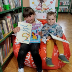 W bibliotece. Dziewczynka i chłopiec siedzą na pomarańczowej pufie. Trzymają dyplomy. W tle regały z książkami.