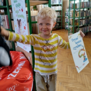 W bibliotece. Uśmiechnięty chłopiec trzyma dyplom i  pluszową maskotkę. W tle regały z książkami.