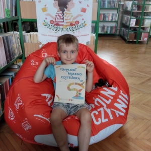 W pomieszczeniu biblioteki. Chłopiec siedzi na pomarańczowej pufie. W ręku trzyma dyplom. W tle regały z książkami.