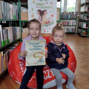 W bibliotece. Dwie dziewczynki siedzą na pomarańczowej pufie. W tle regały z książkami.