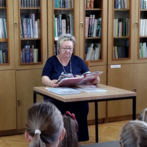 W bibliotece. Przy stoliku siedzi kobieta, która trzyma książkę w rękach.