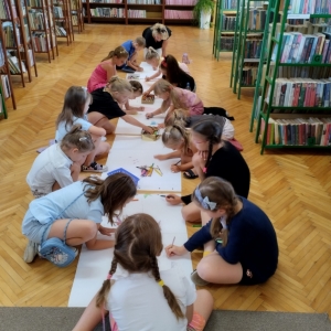 W bibliotece. Na podłodze rozłożone białe kartki. Wokół nich siedzą dzieci i wykonują rysunki.