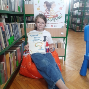 W pomieszczeniu biblioteki. Dziewczynka siedzi na pomarańczowej pufie. W ręku trzyma dyplom. W tle regały z książkami.