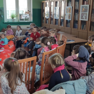 W budynku biblioteki. Grupa dzieci siedzi na pufach i krzesłach. W tle widać przeszklone szafy, regał z książkami i scenę przygotowaną do teatrzyku