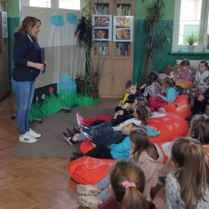 W budynku biblioteki. Po prawej stronie grupa dzieci, siedzących na pufach i krzesłach. Po lewej stronie stoi pani, kórej dzieci słuchają. W tle przeszklony regał z książkami i kotara