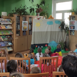 W budynku biblioteki. Grupa dzieci siedzi i patrzy na stojącą przed nimi kotarę. Na kotarze przyczepione chmury, słońce i trawa. Nad kotarą dwie pacynki. Za kotarą widoczne okno. Po lewej i prawej stronie kotary stoją przeszklone szafy z książkami.