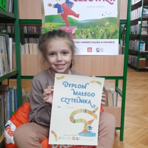 W pomieszczeniu biblioteki. Uśmiechnięta dziewczynka siedzi na pomarańczowej pufie. W ręku trzyma dyplom. W tle regały z książkami.