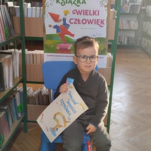 W pomieszczeniu biblioteki. Chłopiec siedzi na niebieskim krześle. W ręku trzyma dyplom.