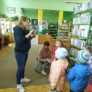 W budynku. Kilkoro dzieci stoi i patrzy na stojącą przed nimi kobietę. Kobieta pokazuje dzieciom małą karteczkę. W tle regały z książkami, biurko, dwa filary. Na zielonych ścianach obrazy, plakaty i tablice informacyj