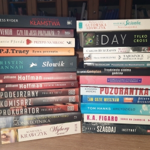 W bibliotece. Na stoliku ułożone dwa stosy książek. W tle regały z książkami.