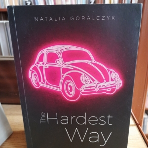 Na stoliku jest ustawiona książka. Książka ma ciemną okładkę, na której zamieszczona jest grafika samochodu w kolorze różowym.