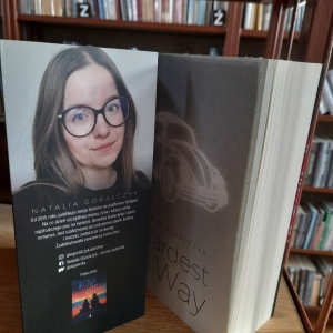 W bibliotece. Na regale ustawiona otwarta książka. W tle widać regały z książkami. 