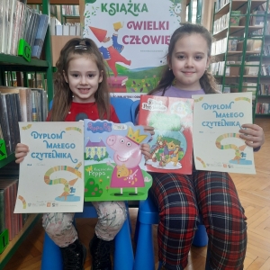 W bibliotece. Dwie dziewczynki siedzą na pomarańczowej pufie. Każda trzyma dyplom i książkę.